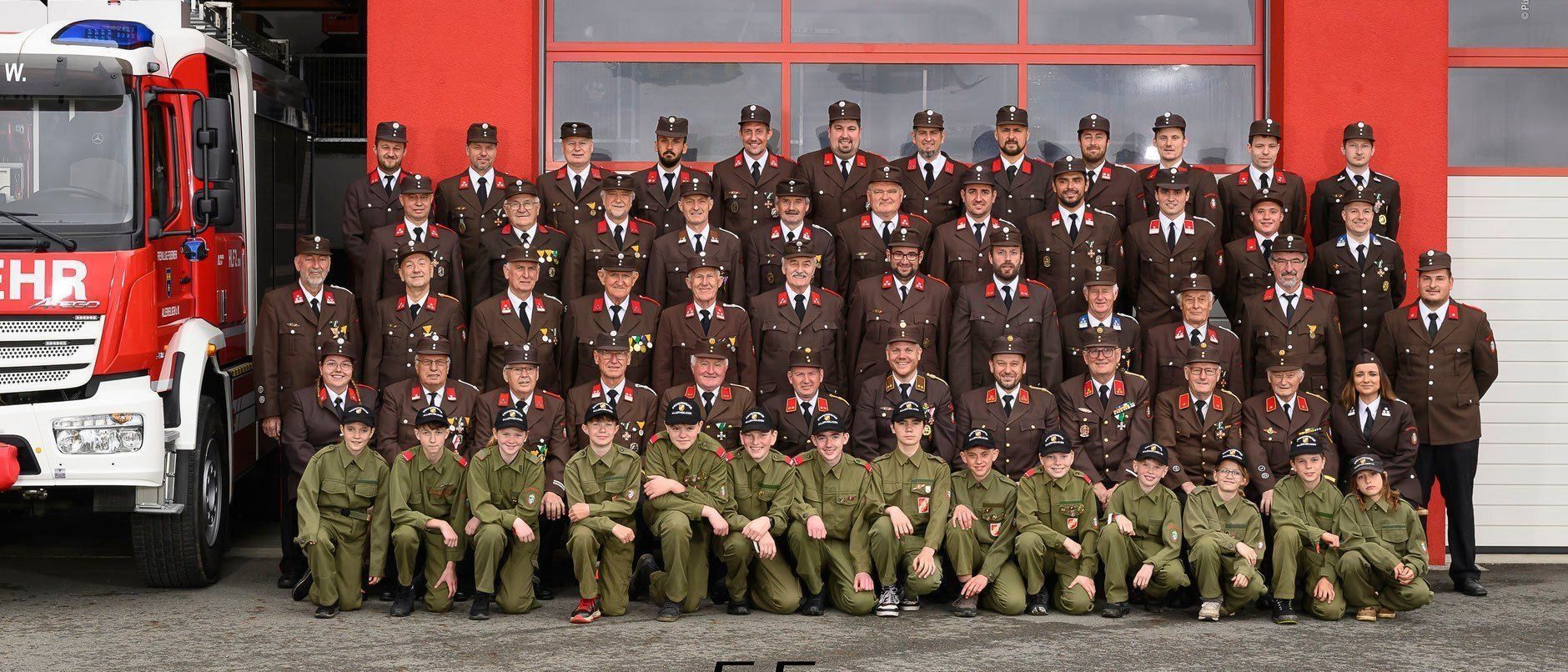 Ein Gruppenfoto von uniformierten Personen steht vor einem roten Gebäude. Sie tragen Mützen und haben Abzeichen an ihren Uniformen. Es gibt zwei Reihen, eine kniend und eine stehend. Der Text lautet '03 Allerheiligen 20'.