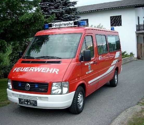 Ein roter Feuerwehrwagen mit weißen Markierungen ist auf einer Einfahrt vor einem weißen Haus mit Balkon geparkt.