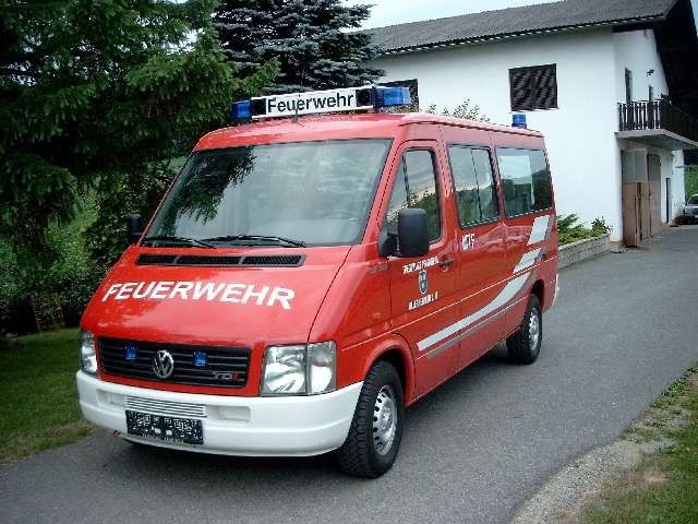 Ein roter Feuerwehrwagen mit weißen Markierungen ist auf einer Einfahrt vor einem weißen Haus mit Balkon geparkt.