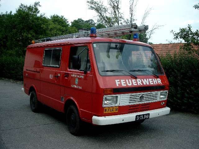 Ein roter VW-Transporter mit einer Leiter auf dem Dach steht auf einem Parkplatz. Auf dem Van steht das Wort 'Feuerwehr'.