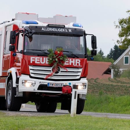 Ein Feuerwehrauto fährt mit Blumen auf der Front, beschriftet mit 'FEUERWEHR'. Ein Fahrer ist im Inneren, mit Häusern und Grün im Hintergrund.