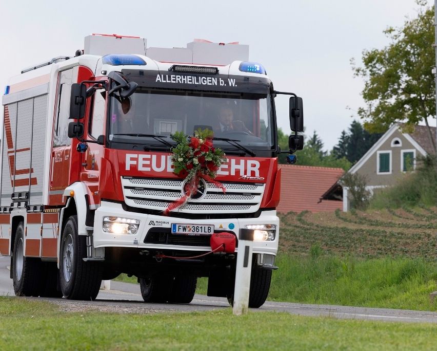Ein Feuerwehrauto fährt mit Blumen auf der Front, beschriftet mit 'FEUERWEHR'. Ein Fahrer ist im Inneren, mit Häusern und Grün im Hintergrund.