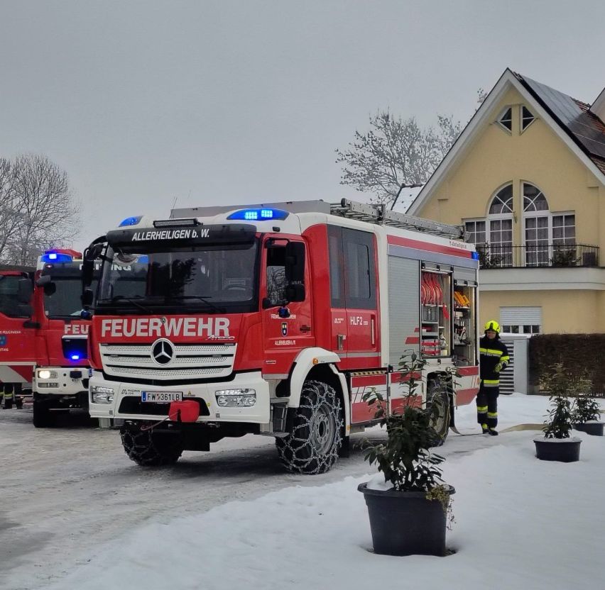 Ein Feuerwehrauto mit der Aufschrift 'Feuerwehr' steht vor einem Haus im Schnee. Ein Feuerwehrmann steht neben dem Auto.