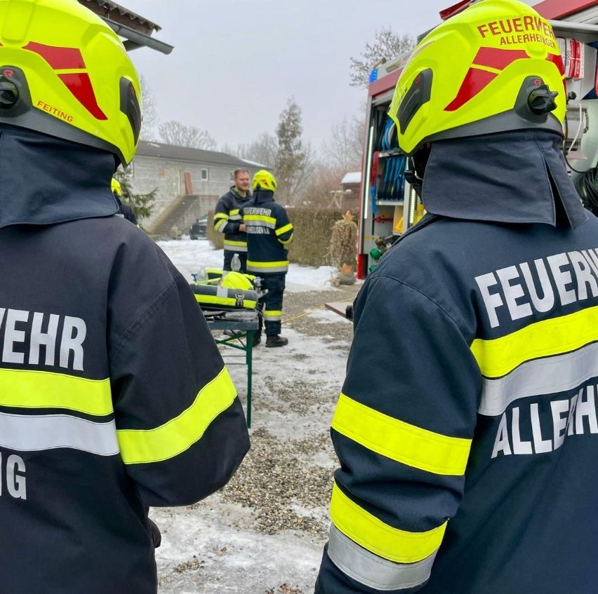 Feuerwehrleute in Schutzkleidung stehen im Schnee, einer vor einem Feuerwehrauto und einer dahinter. Ein dritter Feuerwehrmann ist in der Nähe und führt ein Gespräch.