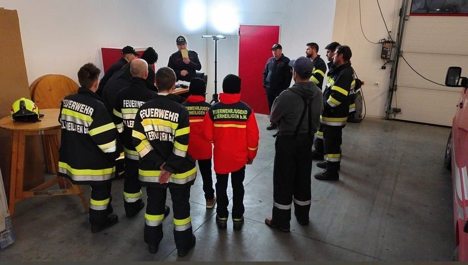Feuerwehrleute stehen in einem Raum, einer vor ihnen hält ein Papier. Hinter ihnen stehen zwei Männer, einer von ihnen spricht.