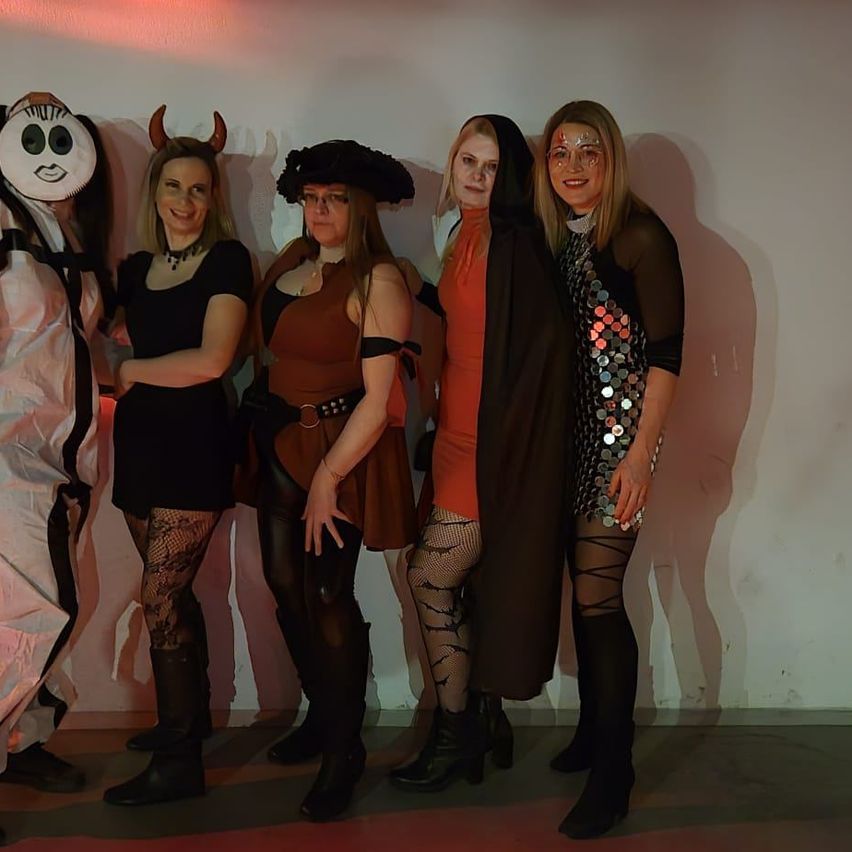Fünf Frauen in Kostümen stehen zusammen und lächeln für ein Foto. Sie tragen verschiedene Kostüme, darunter eine Maske, eine Piratenhaube, einen Teufelshorn und ein Paillettenkleid.