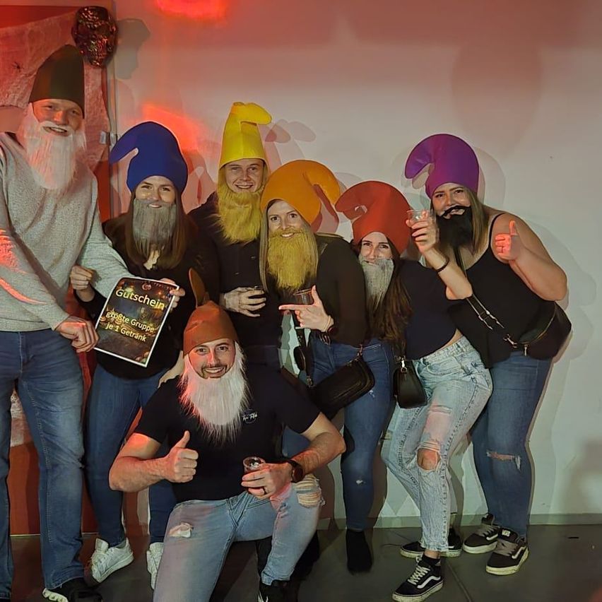Eine Gruppe von Menschen in Zwergenkostümen posiert für ein Foto. Eine Person hält ein Buch, und eine andere gibt einen Daumen hoch. Die Wand ist weiß gestrichen, mit einigen Gegenständen, die hängen.
