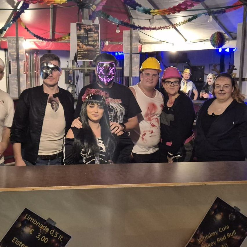 Eine Gruppe von Menschen in Kostümen und Masken posiert für ein Foto in einer Bar mit Getränken und Dekorationen.