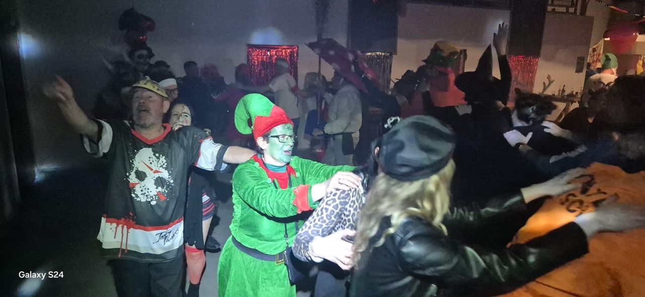 Eine Gruppe von Menschen tanzt auf einer Party; eine Person in einem grünen Elfenkostüm tanzt mit einer anderen Person, die eine Lederjacke trägt.