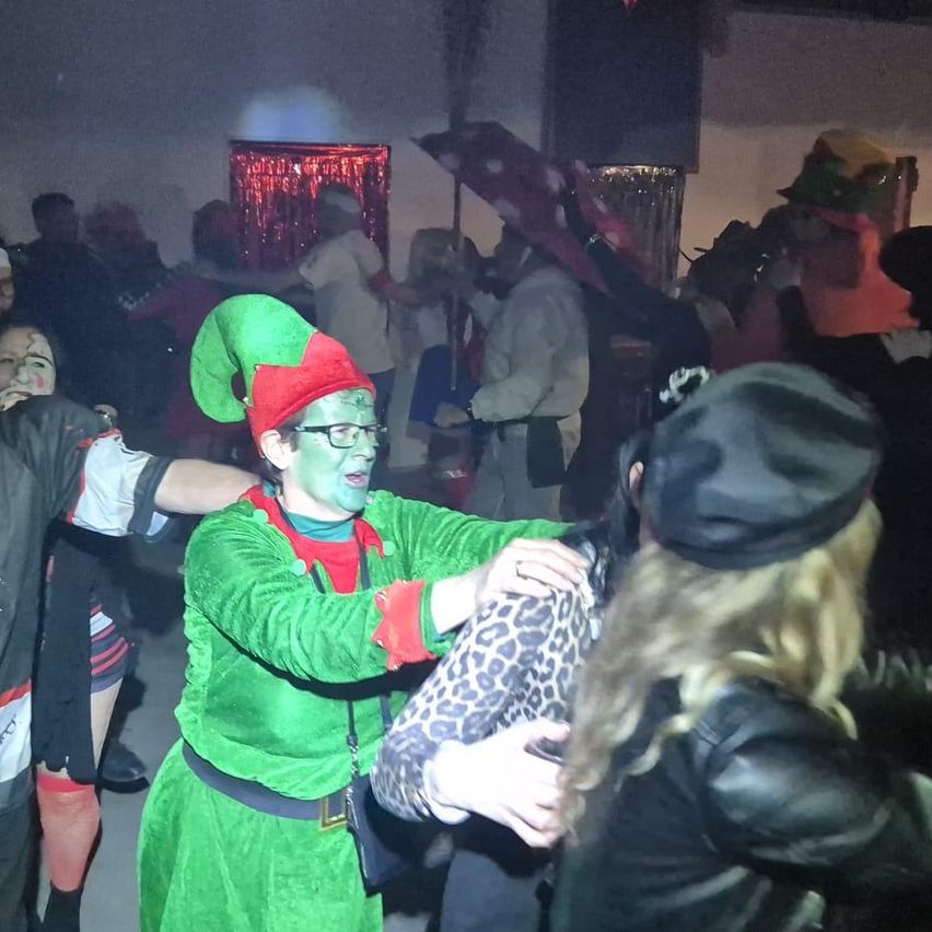 Eine Gruppe von Menschen tanzt auf einer Party; eine Person in einem grünen Elfenkostüm tanzt mit einer anderen Person, die eine Lederjacke trägt.