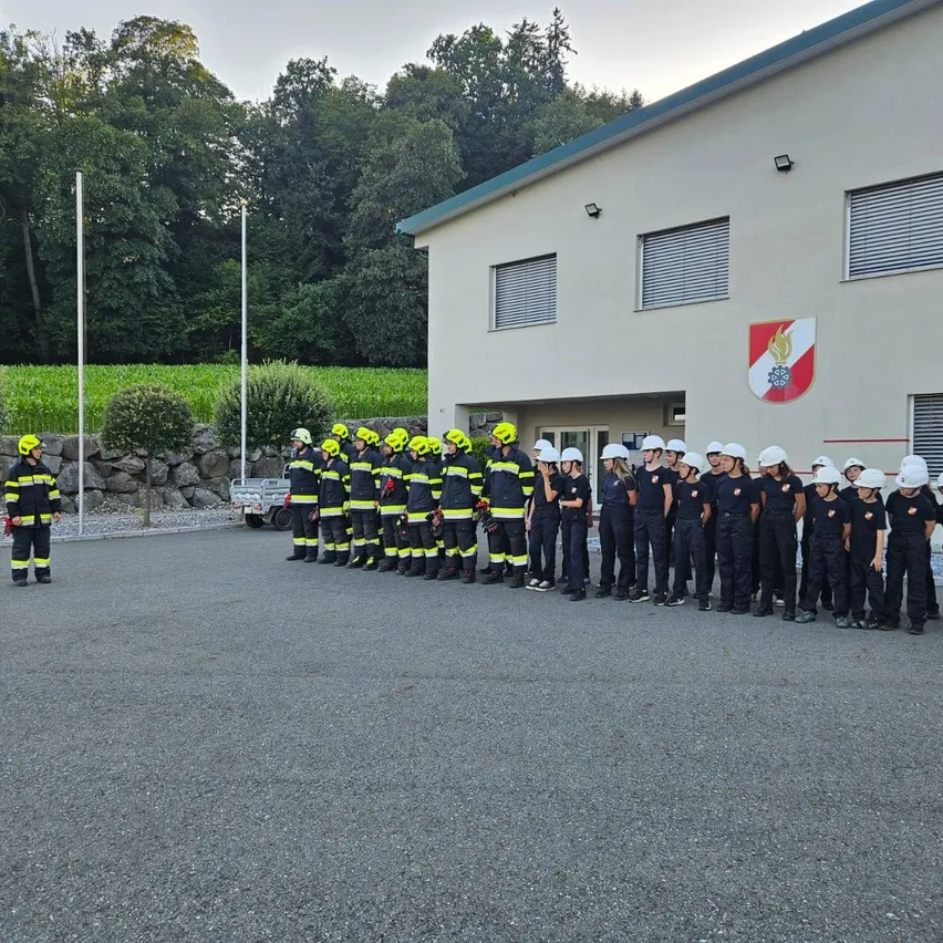 Eine Gruppe von Feuerwehrleuten in Uniform steht vor einem Gebäude mit einem Logo an der Wand.