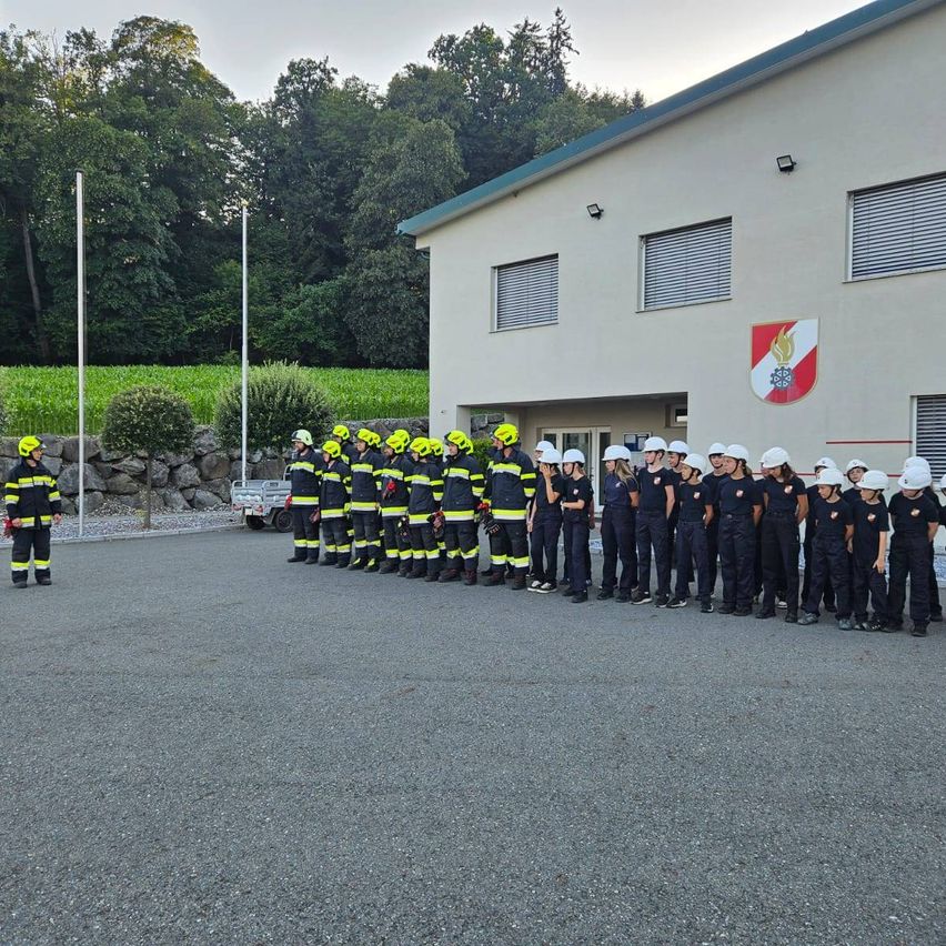 Eine Gruppe von Feuerwehrleuten in Uniform steht vor einem Gebäude mit einem Logo an der Wand.