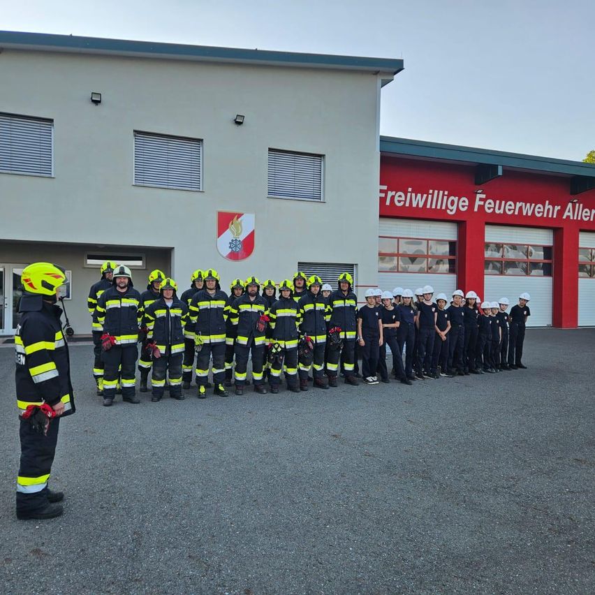 Eine Gruppe Feuerwehrleute in voller Uniform steht vor einem Feuerwehrhaus. Sie tragen Helme und Handschuhe, und das Gebäude hinter ihnen hat ein Logo und Text.