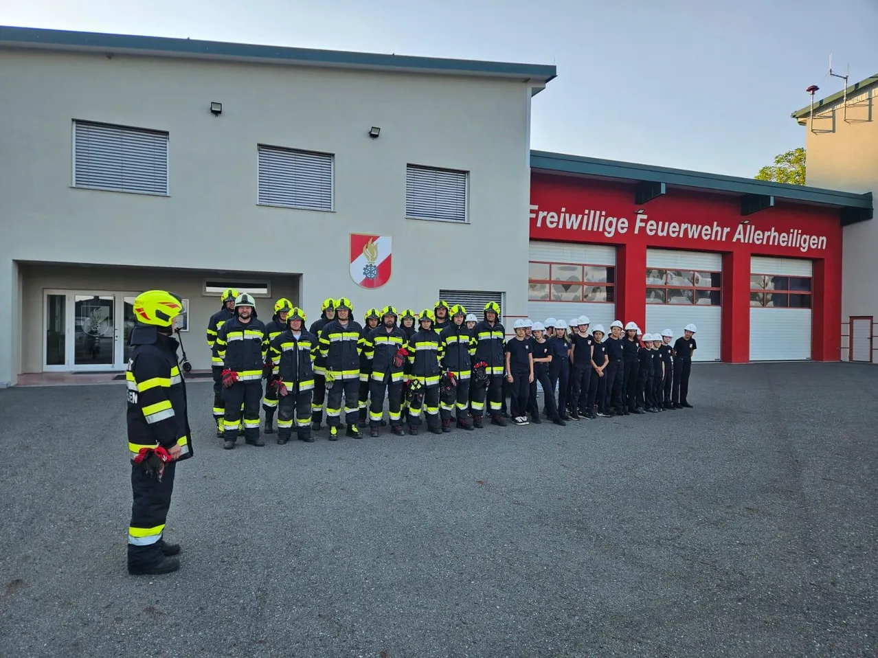 Eine Gruppe Feuerwehrleute in voller Uniform steht vor einem Feuerwehrhaus. Sie tragen Helme und Handschuhe, und das Gebäude hinter ihnen hat ein Logo und Text.