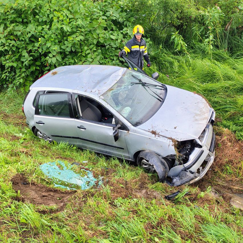 Ein beschädigtes Auto liegt auf der Seite in einem Grasbereich mit einer Person in einem gelben Helm in der Nähe.