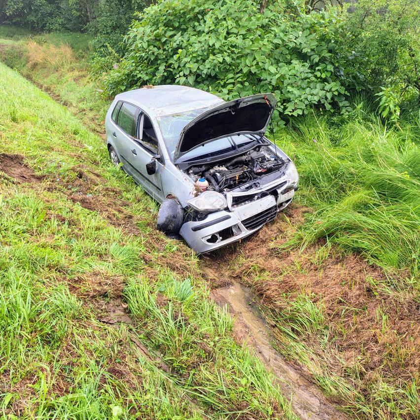 Ein silberner Wagen ist in einem Grasbereich mit geöffneter Motorhaube kaputt. Der Motor ist sichtbar. Das Auto ist seitlich geneigt, und ein Reifen ist abgelöst.