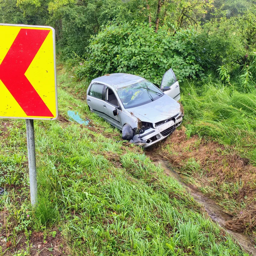 Ein silbernes Auto ist auf einem Grashang beschädigt, mit zertrümmertem vorderen Stoßfänger und offener Fahrertür. Ein gelbes und rotes Schild ist in der Nähe.
