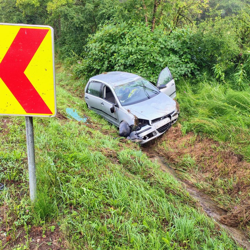 Ein silbernes Auto ist auf einem Grashang beschädigt, mit zertrümmertem vorderen Stoßfänger und offener Fahrertür. Ein gelbes und rotes Schild ist in der Nähe.