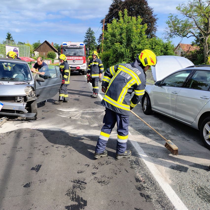Feuerwehrleute in gelben Helmen reinigen die Straße nach einem Autounfall, mit einem beschädigten Auto und einem Feuerwehrwagen im Hintergrund.