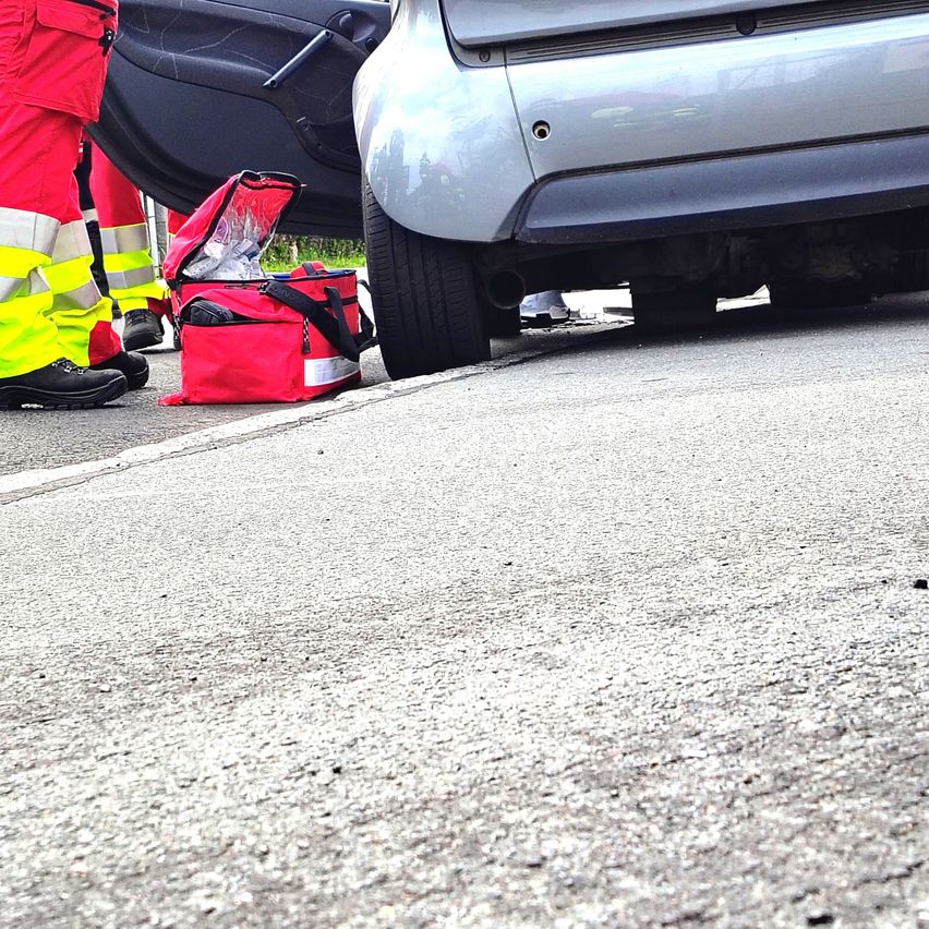 Unfallstelle mit Rettungskräften, die sich um das Fahrzeug kümmern. Ein offener roter Beutel mit medizinischen Utensilien liegt auf dem Boden.