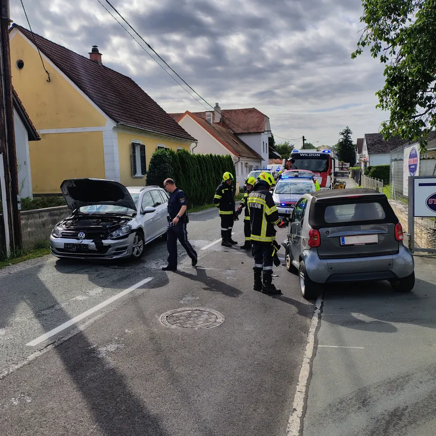 Rettungskräfte kümmern sich um einen Autounfall auf einer Straße. Ein Polizist und Feuerwehrleute sind anwesend, wobei ein Feuerwehrmann das beschädigte Auto untersucht.