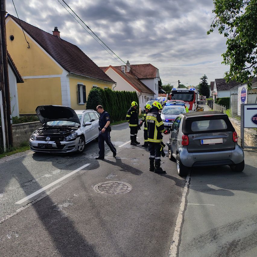 Rettungskräfte kümmern sich um einen Autounfall auf einer Straße. Ein Polizist und Feuerwehrleute sind anwesend, wobei ein Feuerwehrmann das beschädigte Auto untersucht.