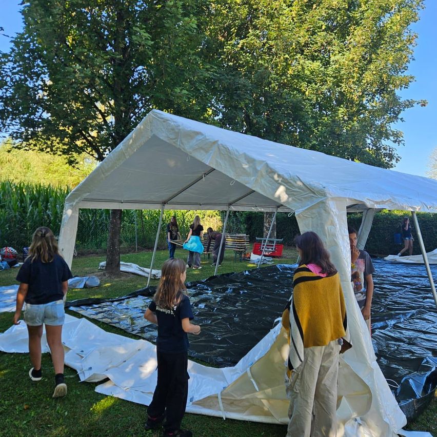 Eine Gruppe von Menschen baut ein weißes Zelt in einem Grasbereich auf, mit Bäumen und Pflanzen im Hintergrund. Einige halten das Zeltgewebe, während andere herumlaufen.