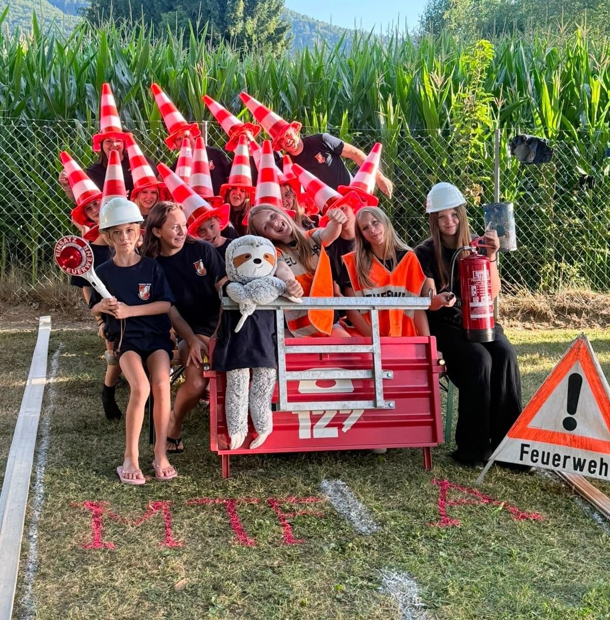 Eine Gruppe von Menschen in Sicherheitswesten und Helmen posiert für ein Foto in einem Maisfeld mit einem Feuerlöscher und einem Kuscheltier auf einer roten Werkzeugkiste.