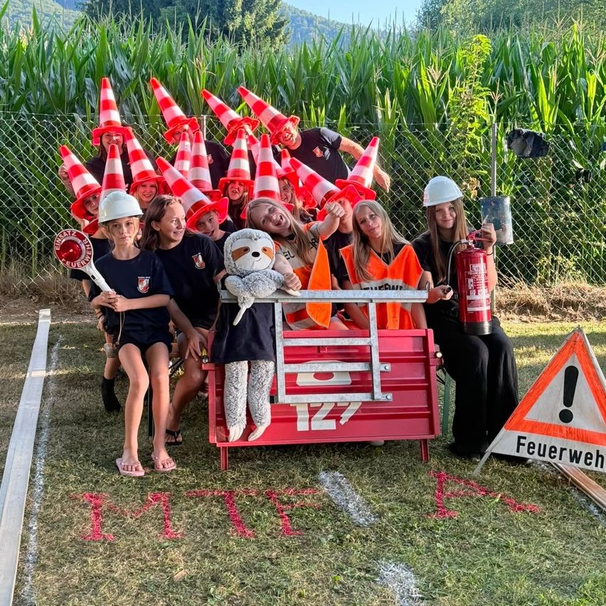 Eine Gruppe von Menschen in Sicherheitswesten und Helmen posiert für ein Foto in einem Maisfeld mit einem Feuerlöscher und einem Kuscheltier auf einer roten Werkzeugkiste.