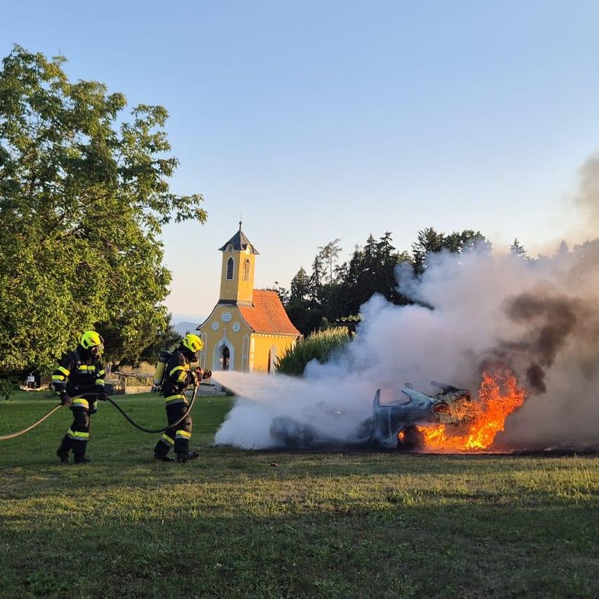 Zwei Feuerwehrleute in voller Ausrüstung löschen ein brennendes Auto vor einer Kirche.