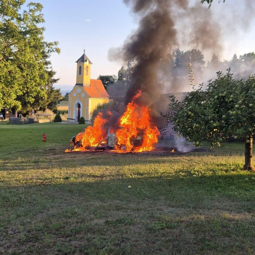 Ein Auto brennt vor einer Kirche mit gelber Fassade und rotem Dach. Bäume und Gras umgeben das Gebiet.