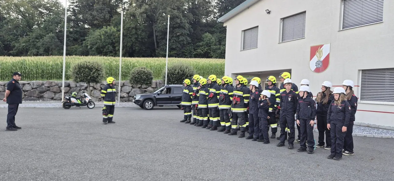 Eine Gruppe von Feuerwehrleuten in gelben Helmen und Uniformen steht in einer Reihe vor einem Gebäude. Ein schwarzer SUV ist in der Nähe geparkt.