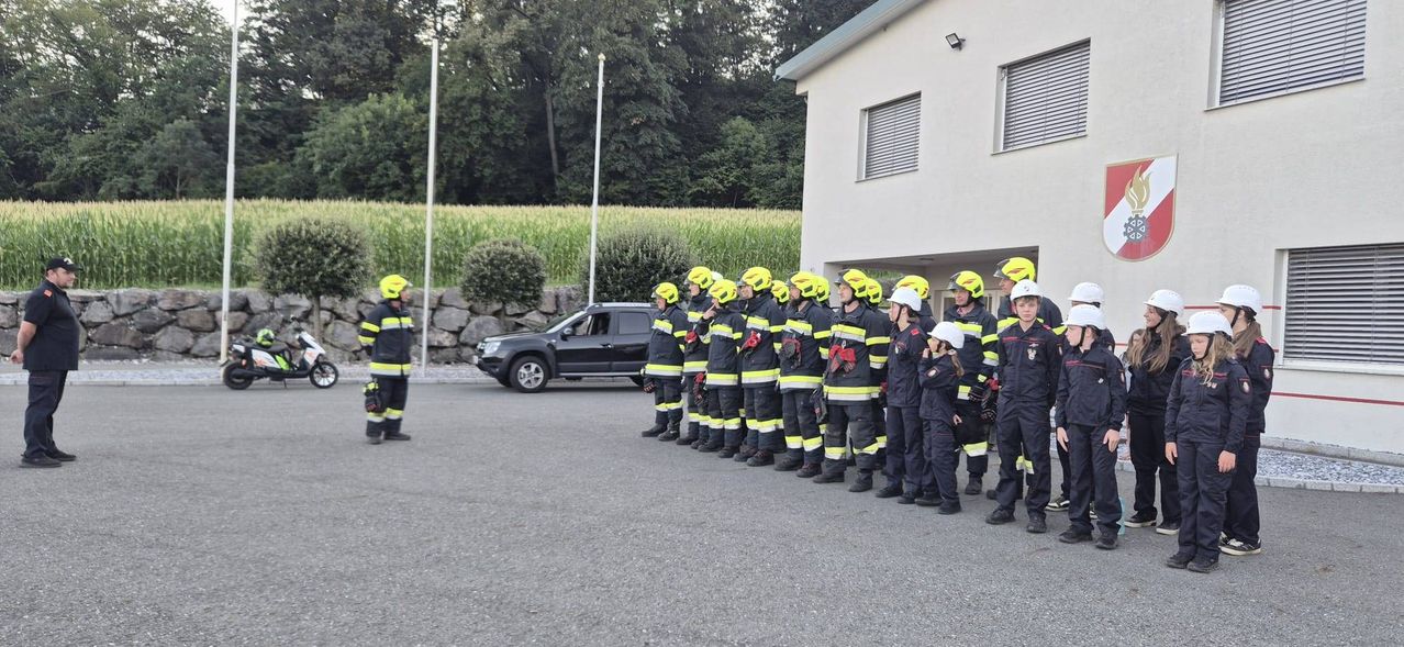 Eine Gruppe von Feuerwehrleuten in gelben Helmen und Uniformen steht in einer Reihe vor einem Gebäude. Ein schwarzer SUV ist in der Nähe geparkt.