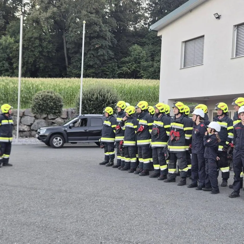 Eine Gruppe von Feuerwehrleuten in gelben Helmen und Uniformen steht in einer Reihe vor einem Gebäude. Ein schwarzer SUV ist in der Nähe geparkt.