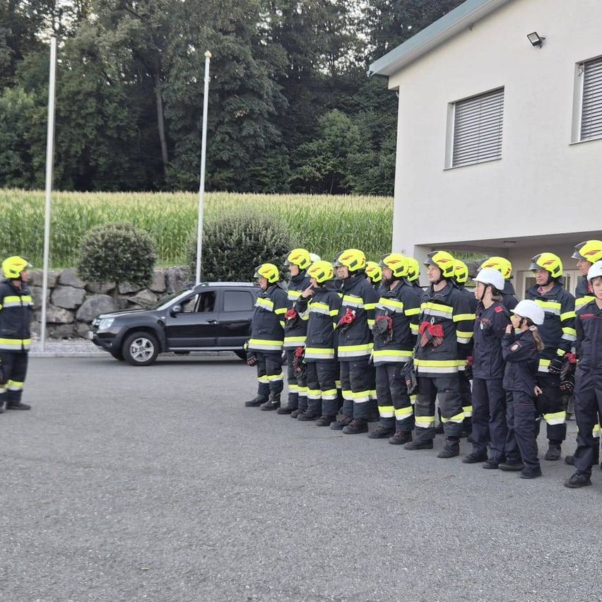 Eine Gruppe von Feuerwehrleuten in gelben Helmen und Uniformen steht in einer Reihe vor einem Gebäude. Ein schwarzer SUV ist in der Nähe geparkt.