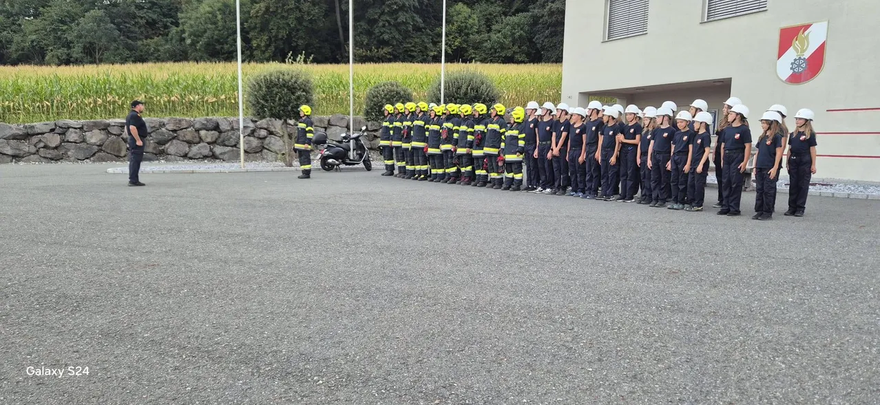Eine Gruppe von Feuerwehrleuten in gelben Helmen und Uniformen steht in einer Reihe vor einem Gebäude. Ein Mann in einem gelben Helm steht links.