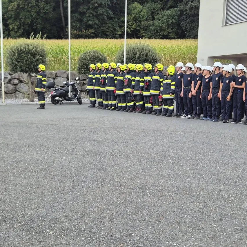 Eine Gruppe von Feuerwehrleuten in gelben Helmen und Uniformen steht in einer Reihe vor einem Gebäude. Ein Mann in einem gelben Helm steht links.