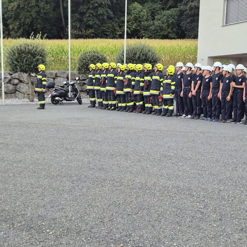 Eine Gruppe von Feuerwehrleuten in gelben Helmen und Uniformen steht in einer Reihe vor einem Gebäude. Ein Mann in einem gelben Helm steht links.