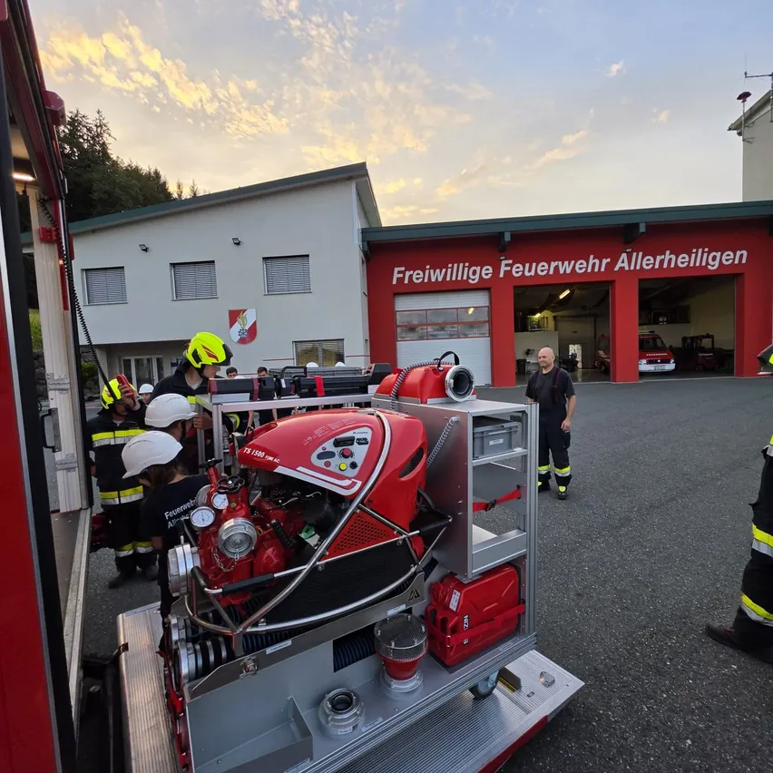 Freiwillige Feuerwehrleute überprüfen einen roten Feuerwehrwagen mit einem großen Motor vor einer Feuerwache.