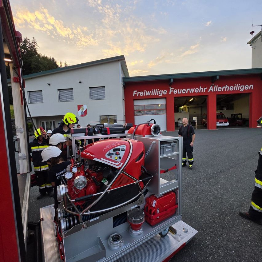 Freiwillige Feuerwehrleute überprüfen einen roten Feuerwehrwagen mit einem großen Motor vor einer Feuerwache.