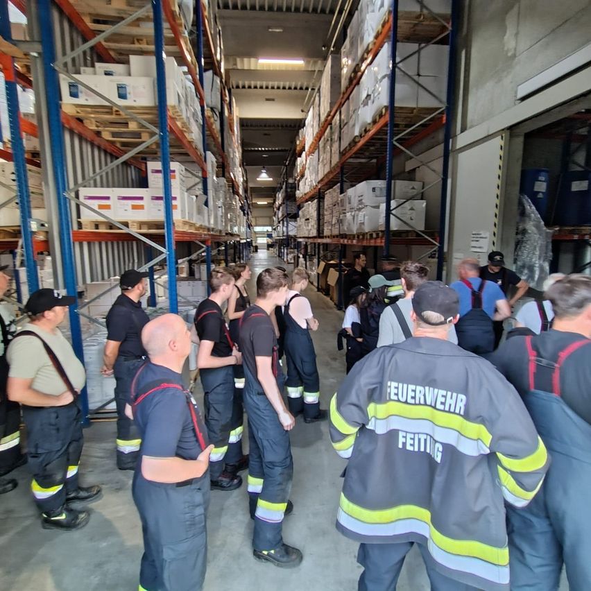 Eine Gruppe Feuerwehrleute in Uniform steht in einem Lagerhaus. Sie sind möglicherweise auf einer Tour, und ein Mann mit schwarzem Hut spricht.