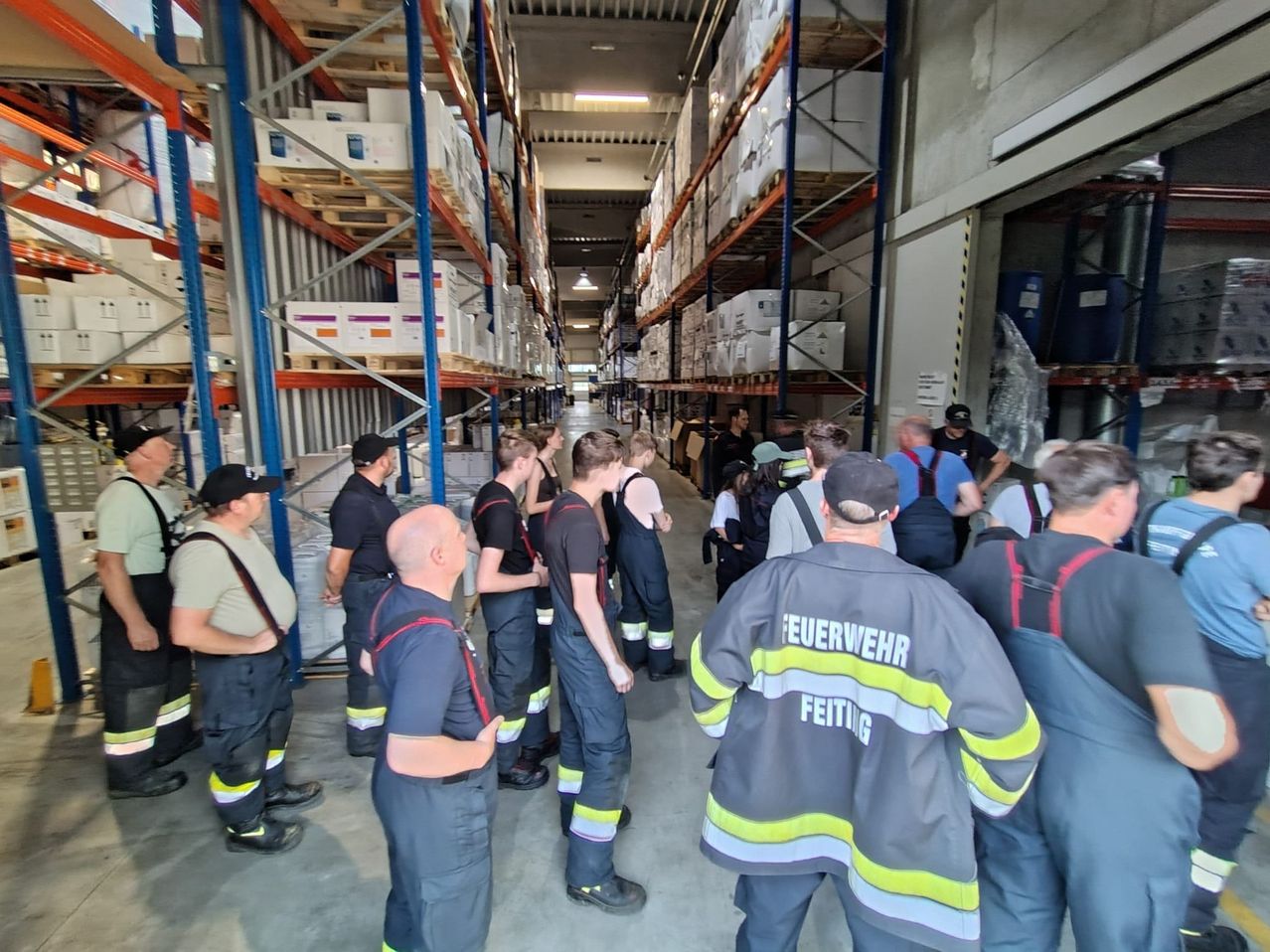 Eine Gruppe Feuerwehrleute in Uniform steht in einem Lagerhaus. Sie sind möglicherweise auf einer Tour, und ein Mann mit schwarzem Hut spricht.