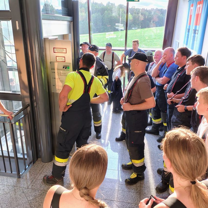 Eine Gruppe von Feuerwehrleuten in Uniformen und Mützen steht in einem Raum, möglicherweise einem Schulungsbereich, mit einer Frau, die von hinten zusieht.