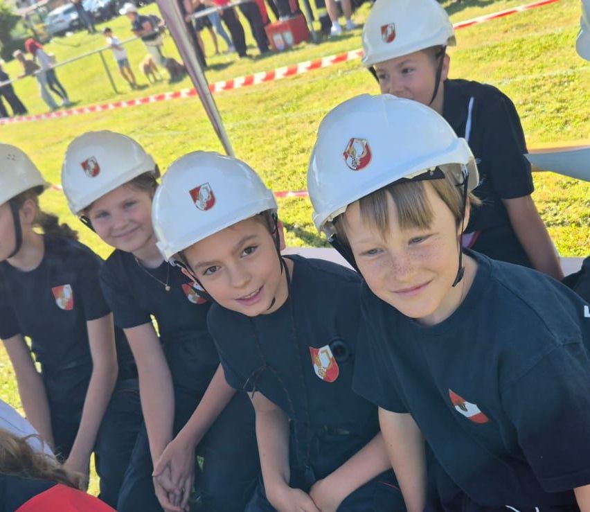 Fünf Kinder tragen weiße Helme mit roten und weißen Logos und sitzen auf einem Grasfeld mit Menschen und Hunden im Hintergrund.