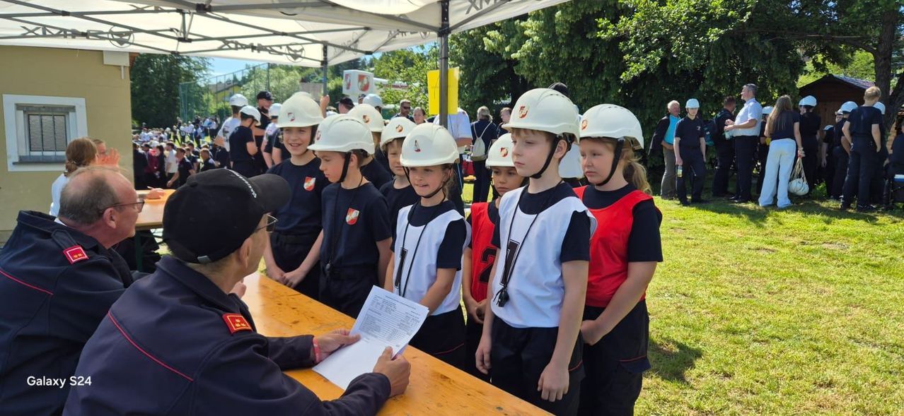Eine Gruppe von Kindern in Helmen steht hinter einem Tisch, an dem ein Mann ein Dokument liest. Bäume und ein Zelt sind im Hintergrund zu sehen.