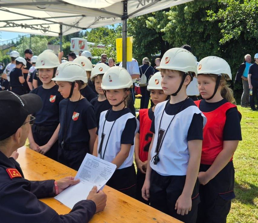 Eine Gruppe von Kindern in Helmen steht hinter einem Tisch, an dem ein Mann ein Dokument liest. Bäume und ein Zelt sind im Hintergrund zu sehen.