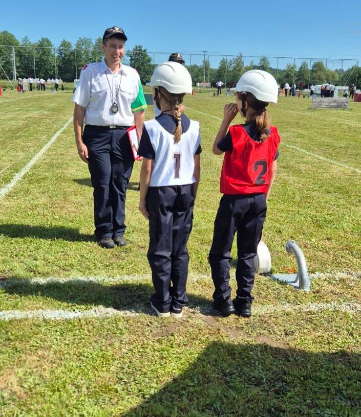 Zwei Kinder in nummerierten Uniformen stehen sich gegenüber, während eine Frau in Uniform hinter ihnen auf einem Feld steht. Bäume und ein Zaun sind im Hintergrund.