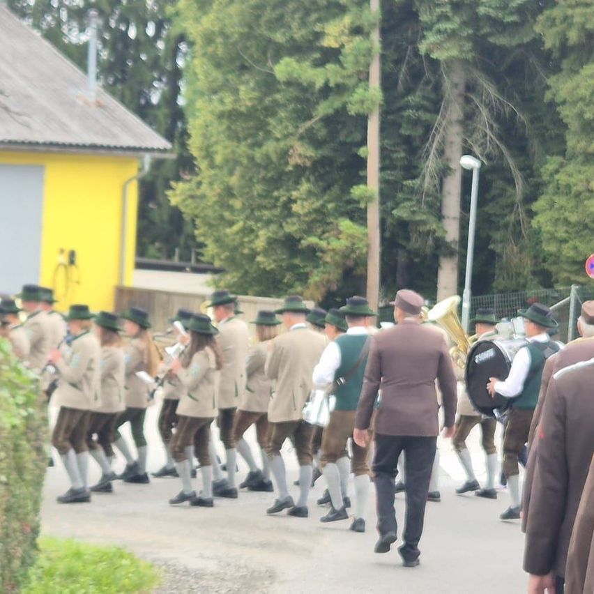 Eine Marschkapelle in traditioneller Kleidung spielt Instrumente und geht die Straße entlang, mit einem gelben Gebäude und Bäumen im Hintergrund.
