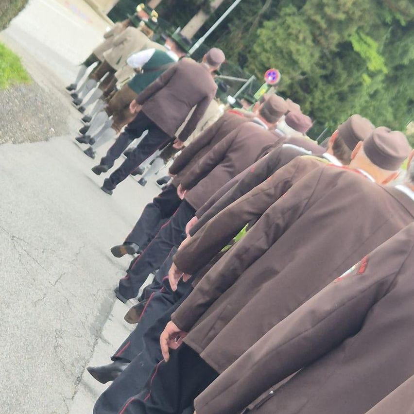 Eine Reihe von Männern in braunen Mänteln und Hüten geht einen gepflasterten Weg hinunter. Im Hintergrund sind Bäume und ein Schild sichtbar.