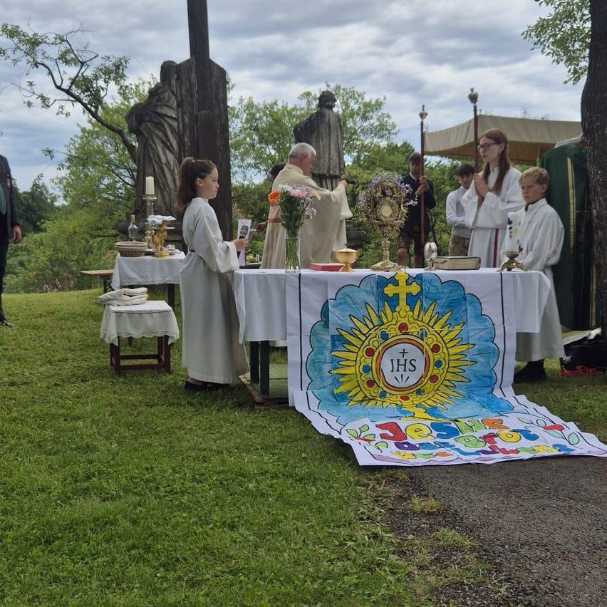 Eine religiöse Zeremonie im Freien mit Altären, einem Priester und Altarjungen. Im Hintergrund befinden sich Statuen und Grünpflanzen.
